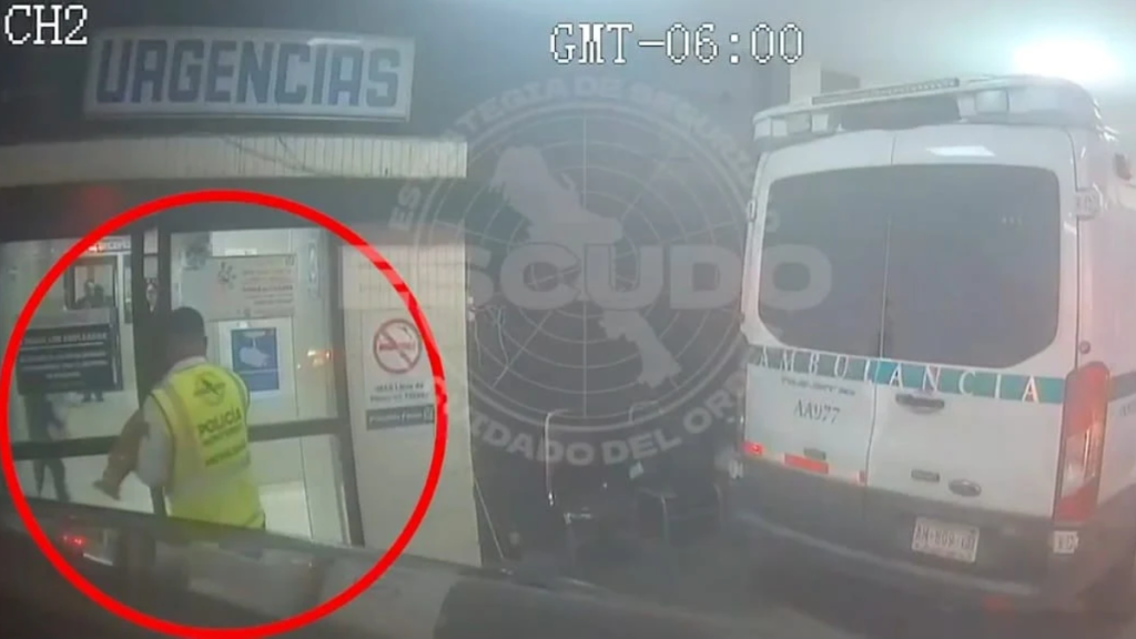 Policeman in a Yellow Reflective Vest Standing at the Entrance of a Building, with an Ambulance Parked to the Right on a Street Scene. - Adelante Mty