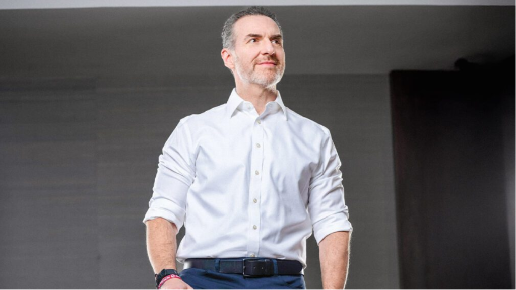 Man in a White Dress Shirt with Rolled Sleeves Stands on Stage, Looking to the Side with a Confident Expression. - Adelante Mty