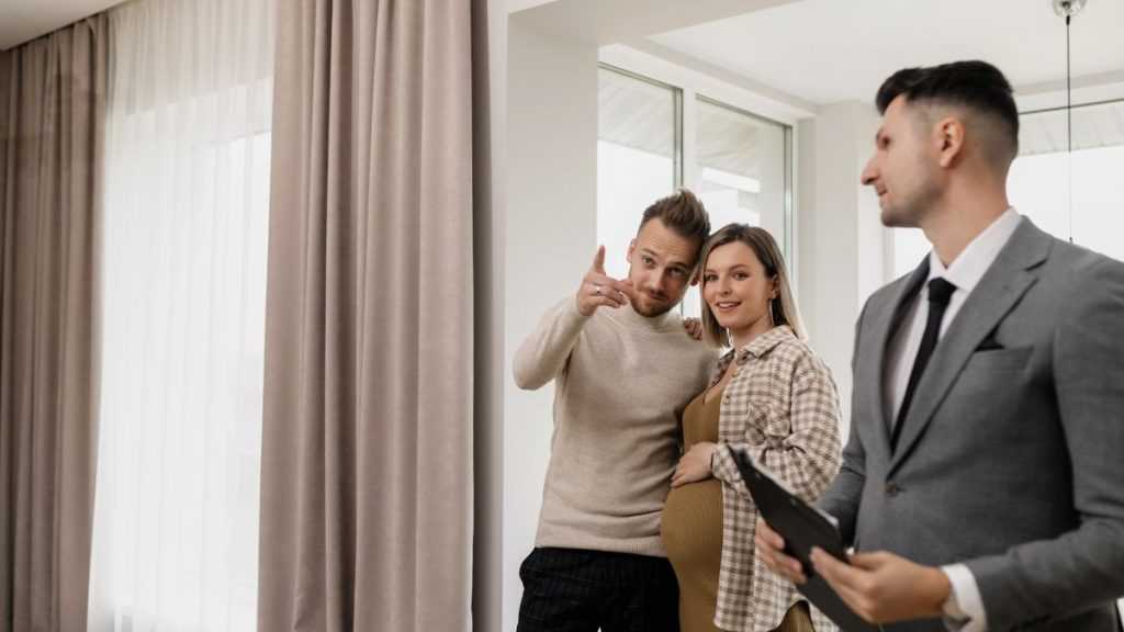 Pregnant Woman and Partner Consult with a Real Estate Agent in a Modern Living Room During a Home Tour, the Man Pointing As They Listen Attentively. - Adelante Mty