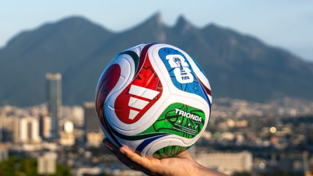 Hand Holding a Colorful Fifa Soccer Ball Outdoors with a Blurred City and Mountain Backdrop. - Adelante Mty