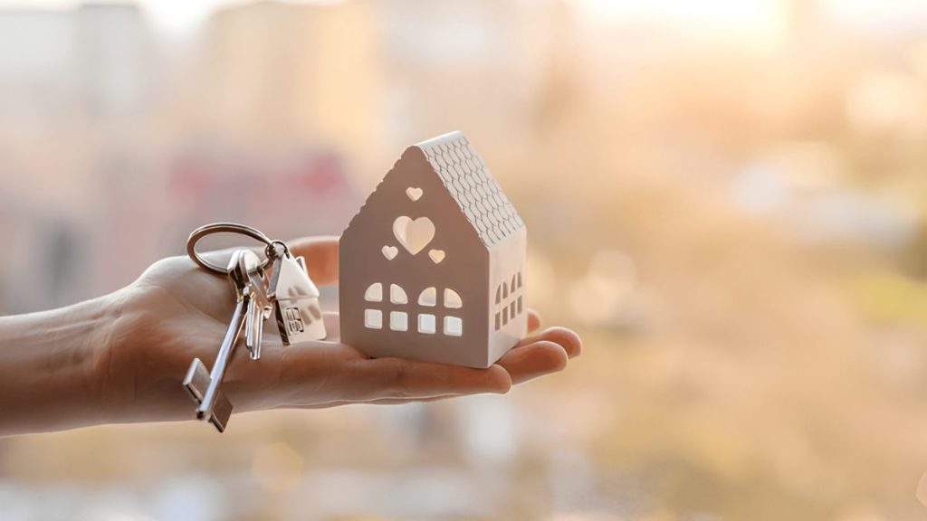Hand Holding a Small Decorative House with a Key Ring, Symbolizing Home Ownership or Real Estate. - Adelante Mty