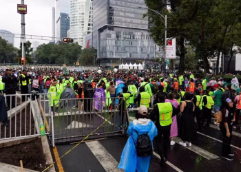 Crowd of People Wearing Bright Safety Vests Gather on a Closed City Street with Tall Modern Buildings in the Background. - Adelante Mty