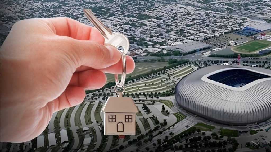 Hand Holding a Key with a Tiny House Charm in Front of an Aerial View of a Stadium and Terraced Landscape. - Adelante Mty