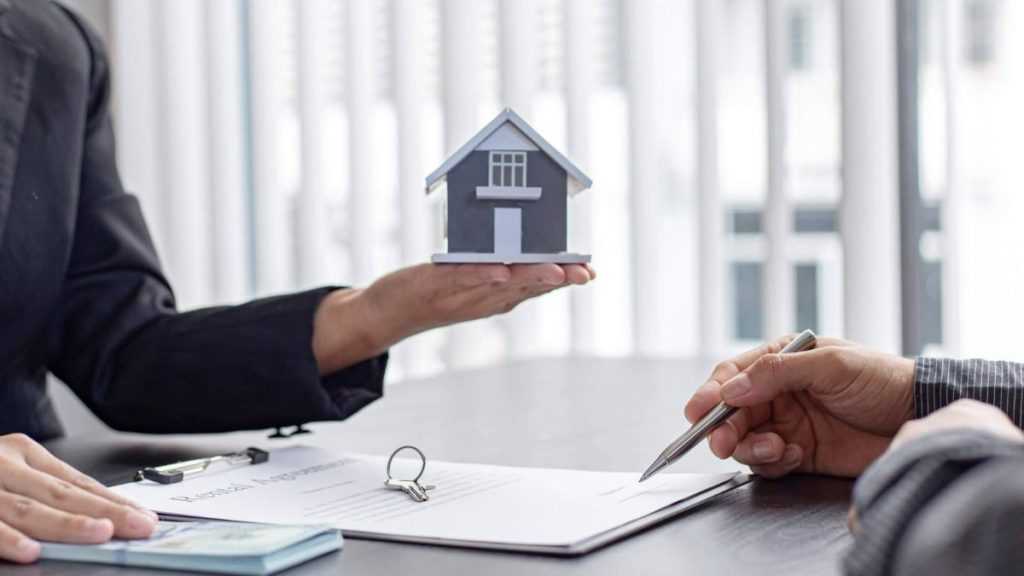 Two People in a Business Setting: One Holds a Small House Model While the Other Signs a Contract or Forms on a Desk - Adelante Mty
