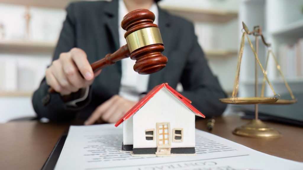 a Judge or Lawyer in a Suit Taps a Wooden Gavel on a Miniature House Placed on Documents, with a Scales of Justice in the Background (legal Real Estate Scene). - Adelante Mty