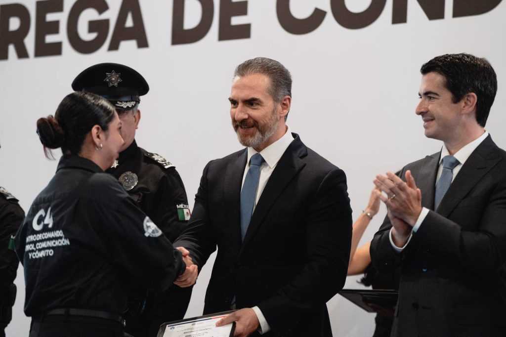 Man in a Suit Shakes Hands with a Female Police Officer As He Presents Her with a Certificate at a Formal Ceremony, While Another Man Congratulates in the Background. - Adelante Mty