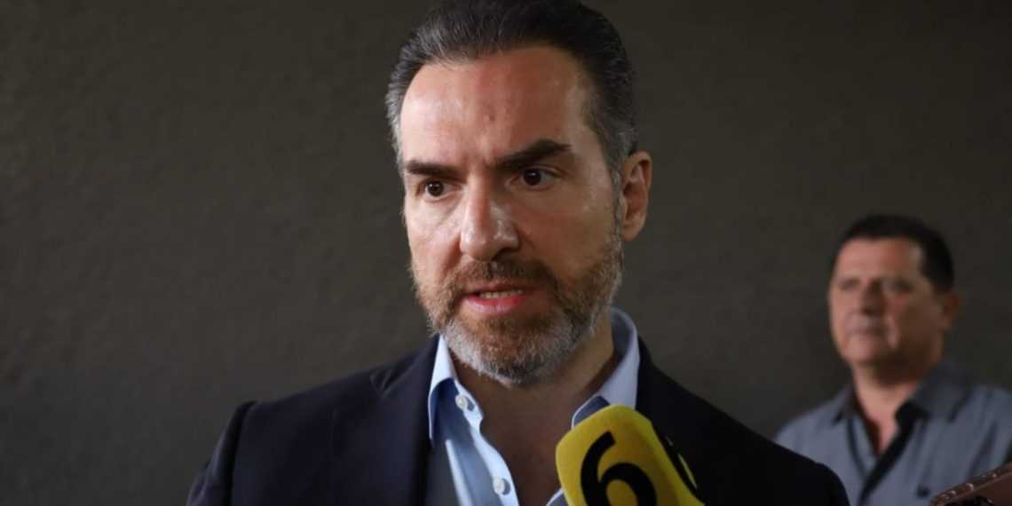 Man in a Dark Blazer Speaks into a Yellow Microphone Labeled '6' at a Press Event, with Another Man in the Background. - Adelante Mty