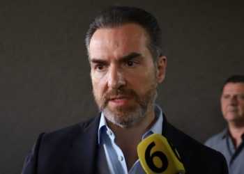 Man in a Dark Blazer Speaks into a Yellow Microphone Labeled '6' at a Press Event, with Another Man in the Background. - Adelante Mty