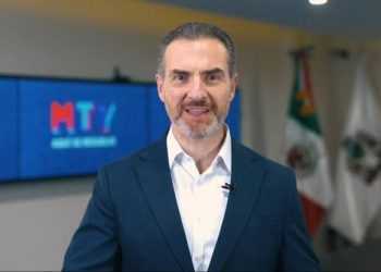 Man in a Dark Suit and White Shirt Speaks to the Camera in a Modern Briefing Room with Large Screens and Flags in the Background. - Adelante Mty