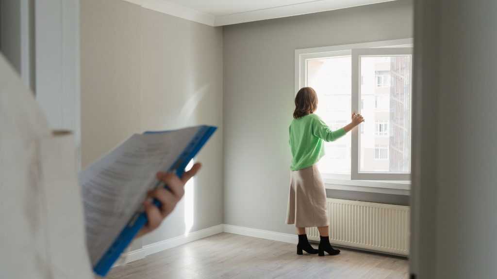 a Person in a Bright Green Sweater Stands by a Window in an Empty Room, While Another Person Holds a Clipboard Nearby. - Adelante Mty
