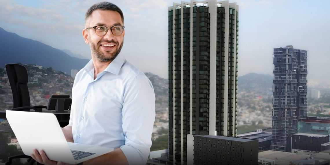 Smiling Man in a Light Blue Shirt Typing on a White Laptop with a City Skyline and Tall Buildings Behind Him in an Office Setting. - Adelante Mty