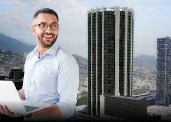 Smiling Man in a Light Blue Shirt Typing on a White Laptop with a City Skyline and Tall Buildings Behind Him in an Office Setting. - Adelante Mty
