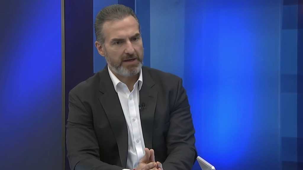 Man in a Dark Suit and White Shirt Seated on a Tv Interview Set with a Blue Gradient Background, Hands Clasped As He Speaks. - Adelante Mty