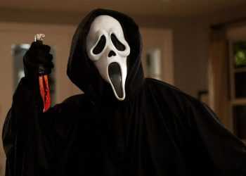 Person in a black hooded cloak wearing a Ghostface mask raises a blood-stained knife in a dimly lit room.