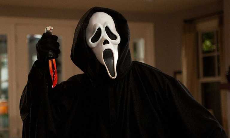 Peliculas-halloween-2021-fleek-1 - Adelante Mty Person in a Black Hooded Cloak Wearing a Ghostface Mask Raises a Blood-stained Knife in a Dimly Lit Room. - Adelante Mty